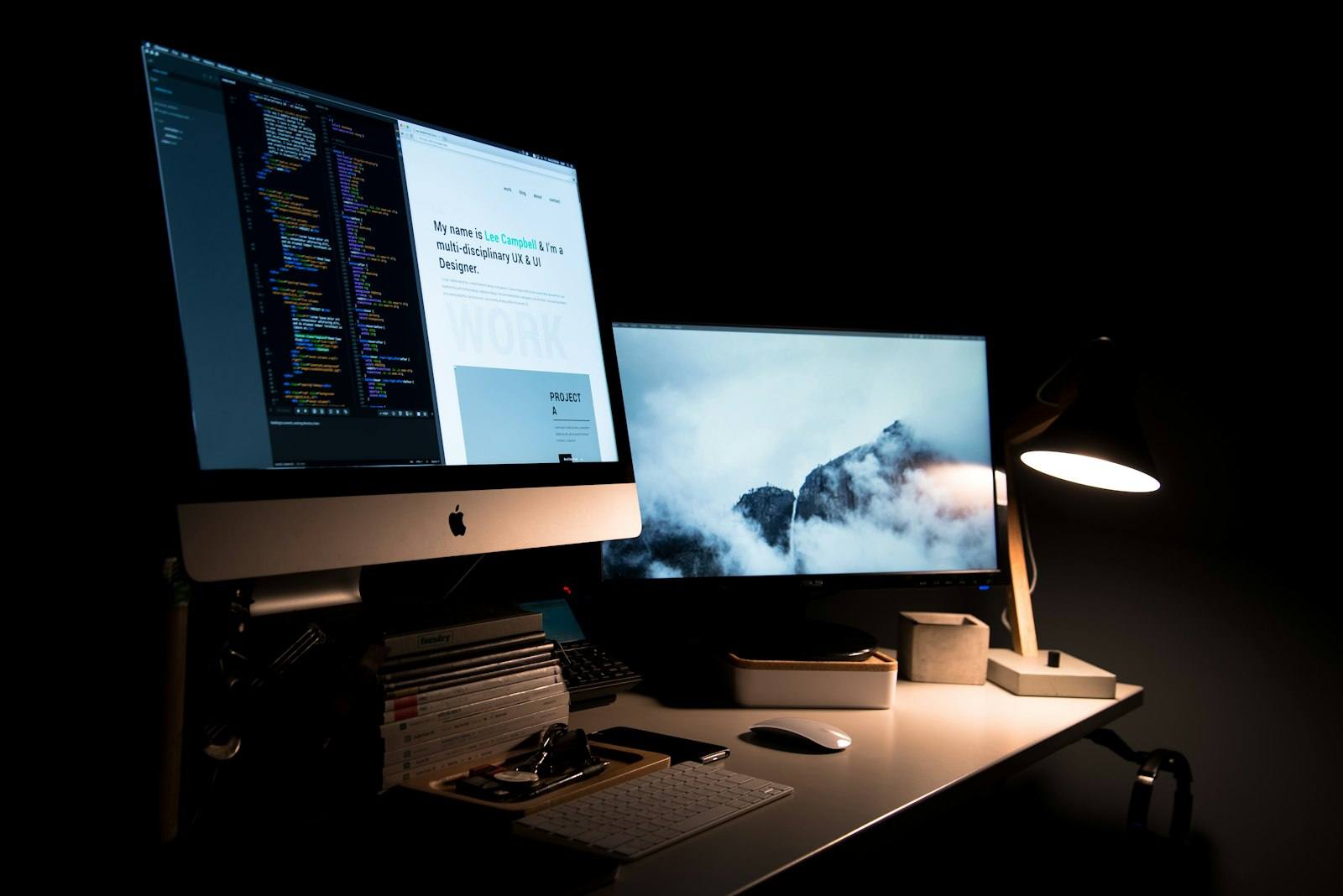 A desktop workstation at night with two monitors showing a website and a lit desk lamp