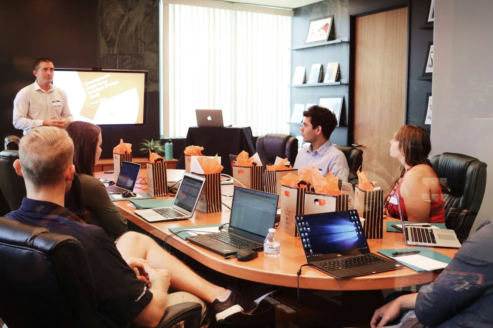 A presenter addressing a seated team in a boardroom with open laptops