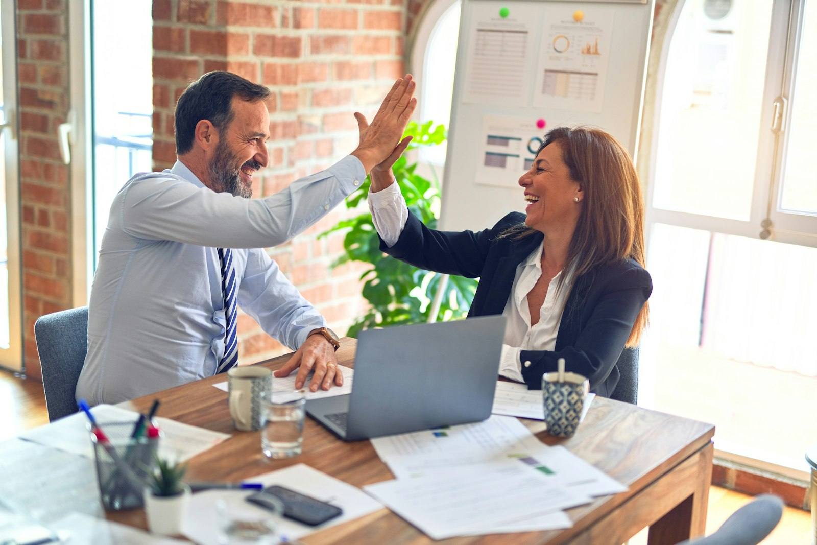 Two colleagues celebrating with a high five at a desk with a laptop